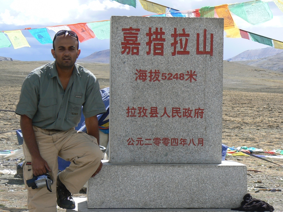 Homme posant à côté d'un monument en pierre avec du texte et des drapeaux de prière.
