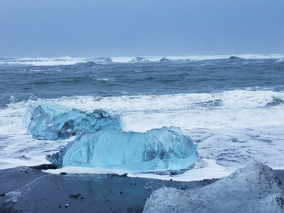 Blocs de glace sur une plage de sable noir avec des vagues qui se brisent en arrière-plan.