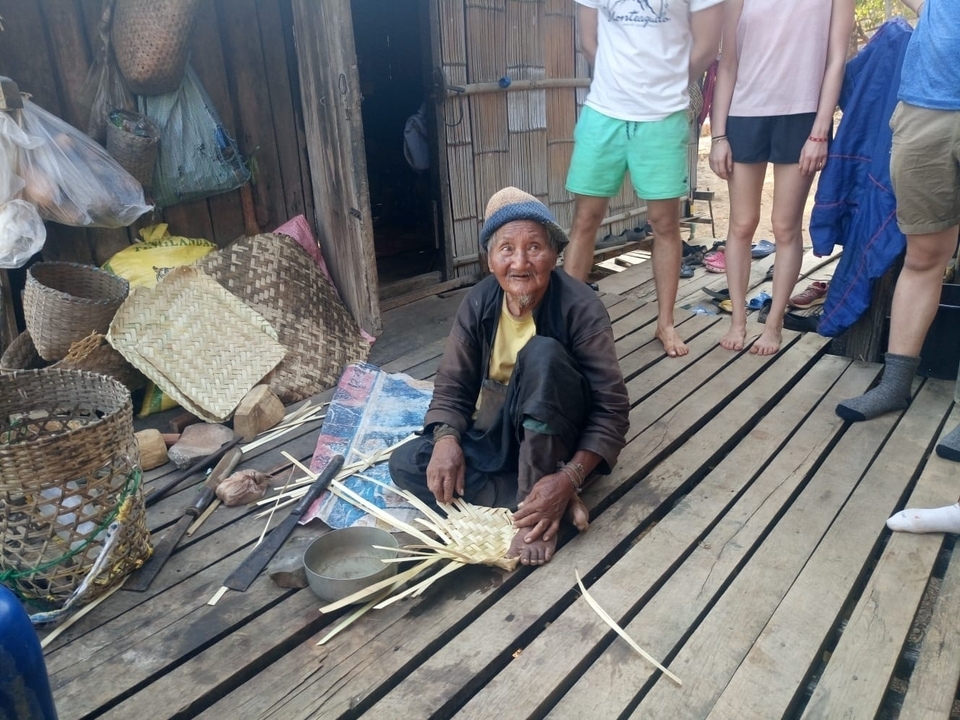 Personne âgée tissant sur un porche avec des gens debout autour.