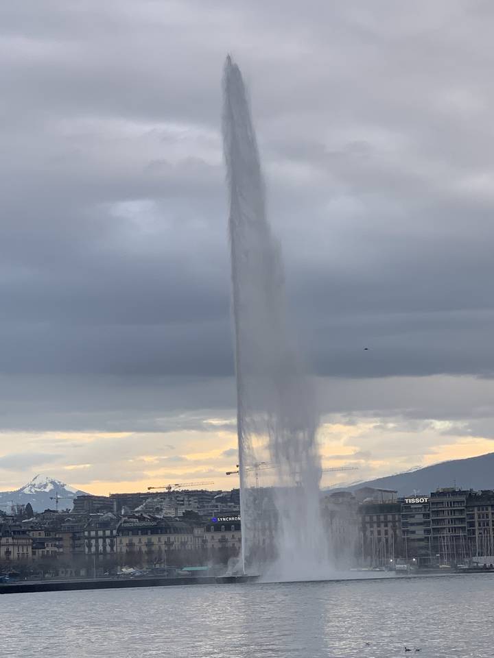 Grand jet de fontaine dans une étendue d'eau, avec un paysage urbain en arrière-plan.