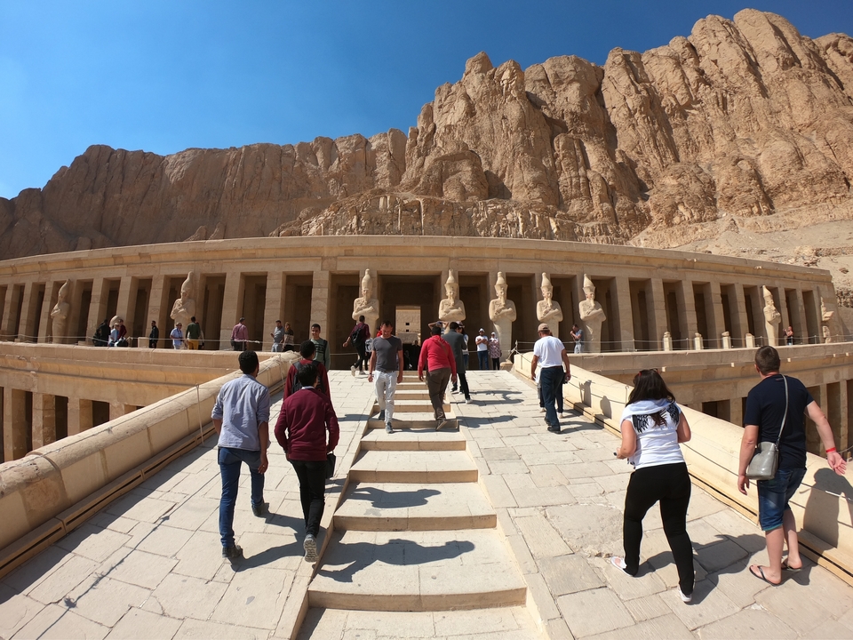 Des visiteurs montant les marches d'un temple égyptien antique avec des falaises rocheuses en arrière-plan.