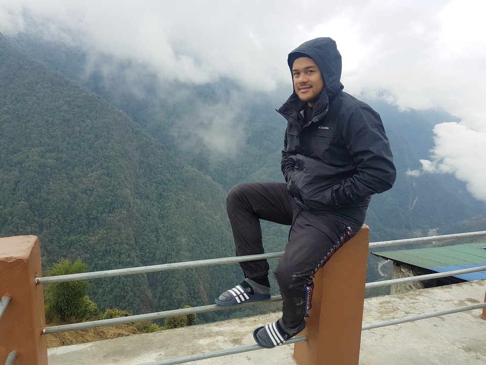 Personne en veste noire assise sur une rambarde surplombant une vallée brumeuse.