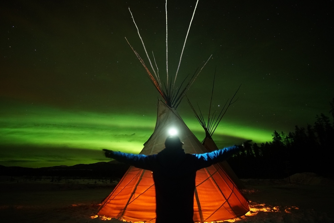 Silhouette d'une personne devant un tipi avec les aurores boréales en arrière-plan.