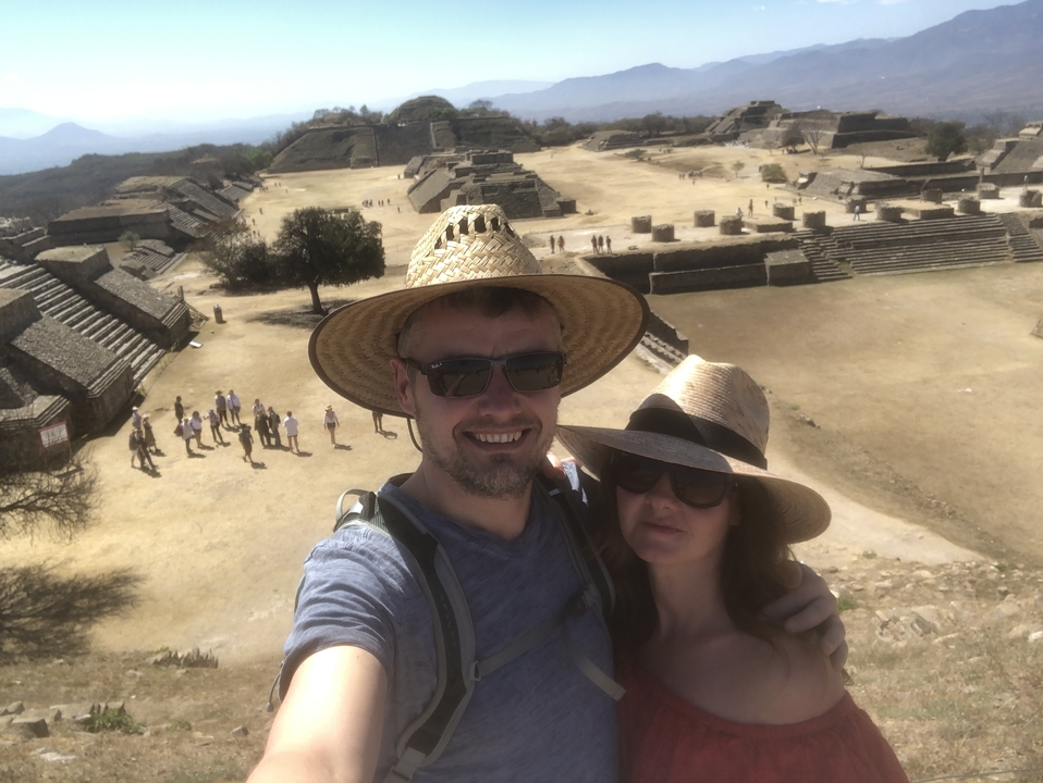 Selfie of a couple in straw hats overlooking the ancient hilltop ruins of Monte Albán under bright sun.