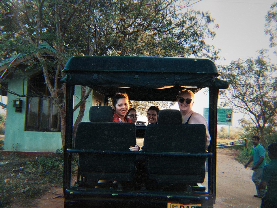 Groupe de personnes dans un véhicule de safari avec des arbres et un petit bâtiment en arrière-plan.