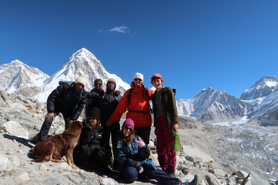 Groupe de personnes posant avec un chien devant des pics montagneux enneigés.