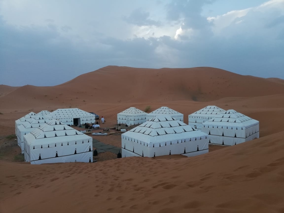 A desert camp with traditional tents surrounded by sand dunes under a cloudy sky.