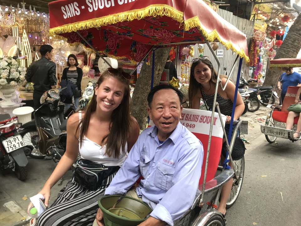 Tourists smiling with a cyclo driver on a bustling Asian street.
