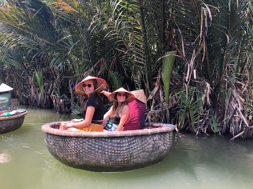 Women wearing conical hats riding a traditional round basket boat through dense palms.