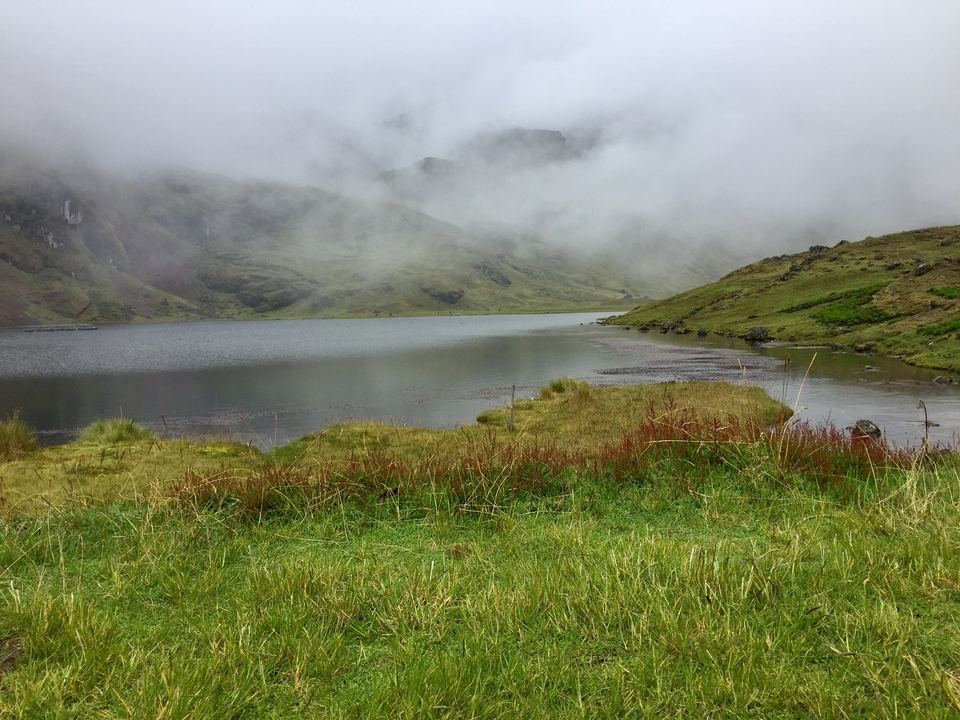 Lac brumeux entouré de collines vertes et de nuages.