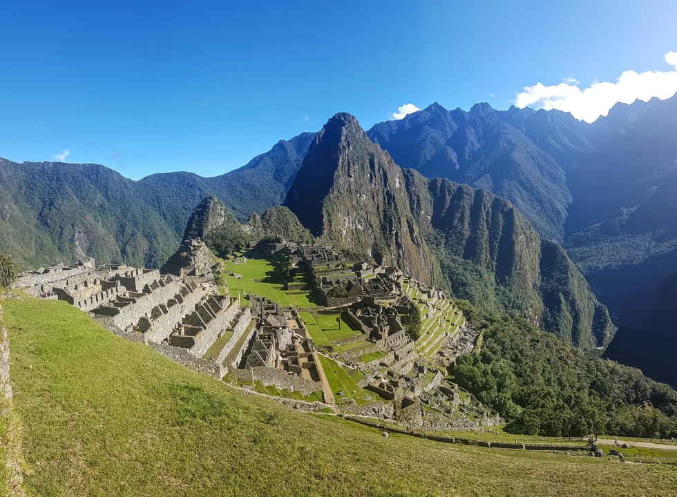 Panoramic postcard view of Machu Picchu ruins and Huayna Picchu peak under clear blue sky.