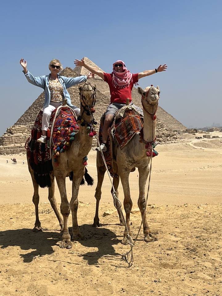 Des gens à dos de chameaux dans un désert avec des pyramides en arrière-plan.