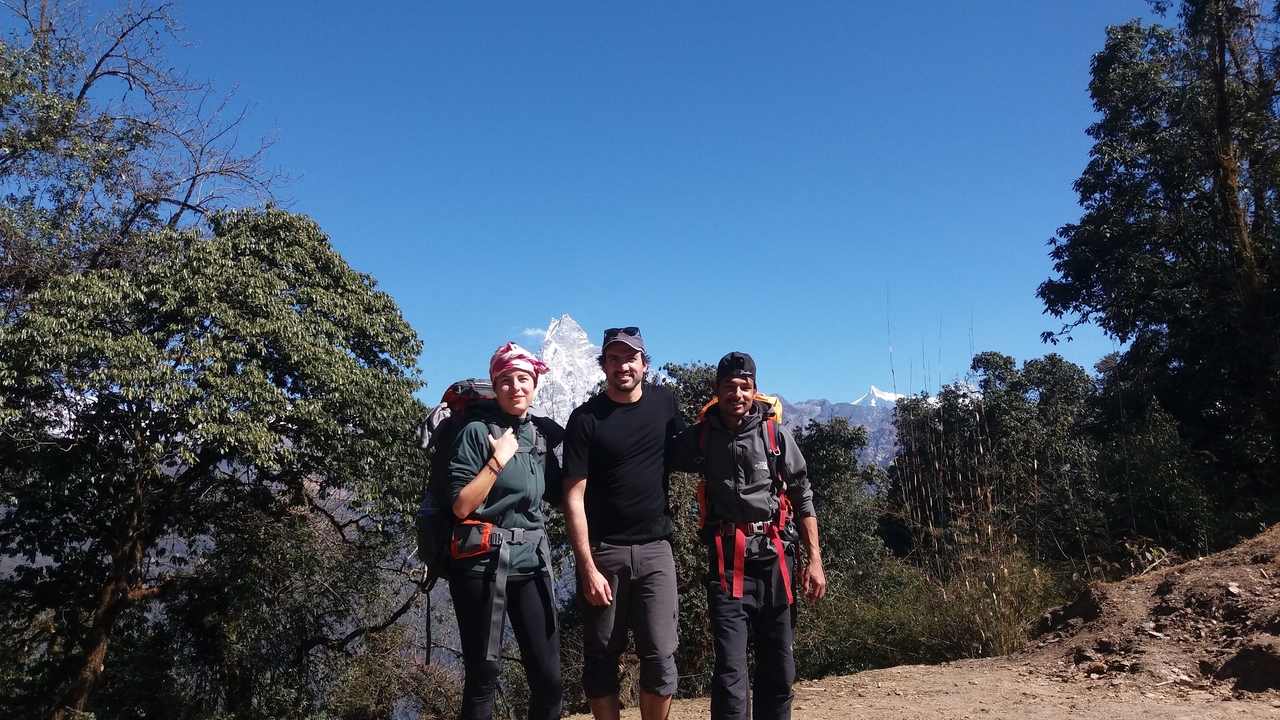 Three trekkers pose on a forested mountain trail with a snow-capped Himalayan peak rising behind them.
