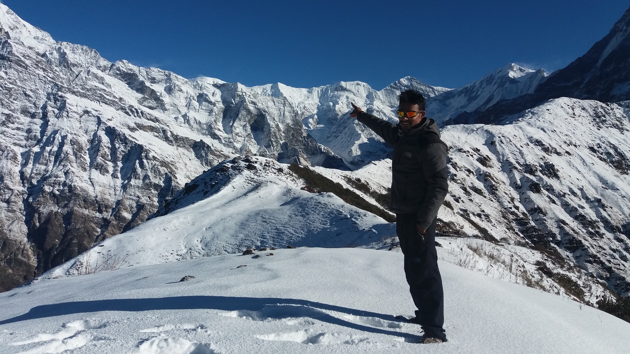 Hiker points toward a jagged snow-covered Himalayan ridge while standing on a white snowy slope under a deep blue sky.