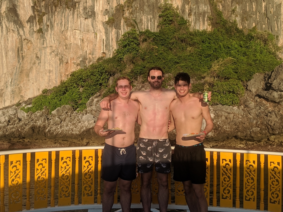 Trois hommes posant torse nu sur un bateau, avec des falaises et de la verdure.