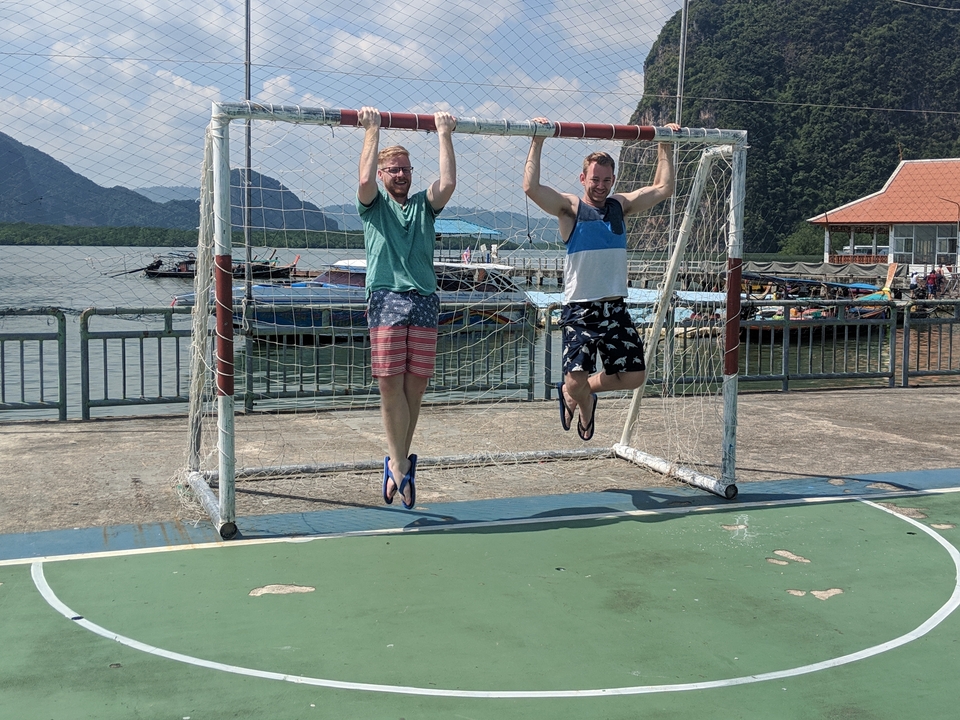 Deux personnes suspendues à un poteau de but de football, avec de l'eau et des montagnes en arrière-plan.