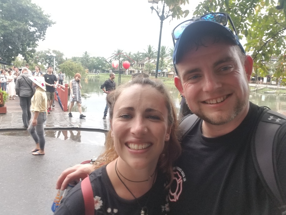 Deux personnes souriantes dans un selfie en extérieur avec un pont en arrière-plan.