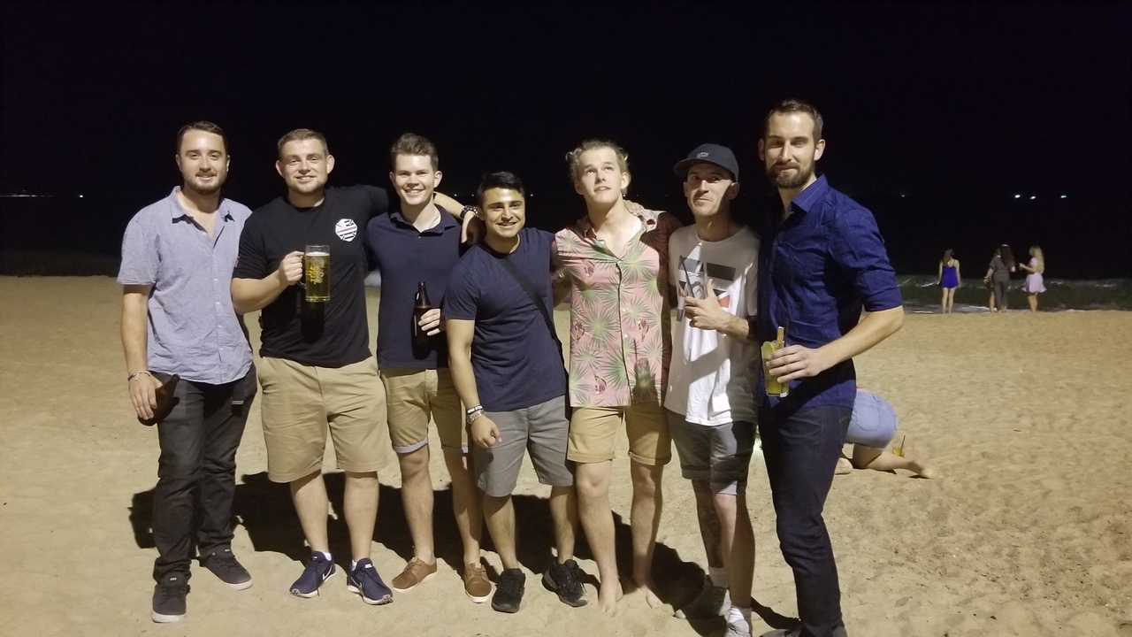 Groupe d'hommes debout sur une plage la nuit, tenant des boissons.