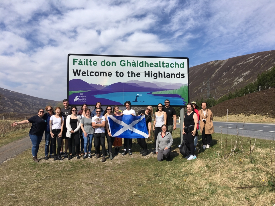 Groupe de personnes tenant un drapeau écossais devant un panneau "Bienvenue dans les Highlands".