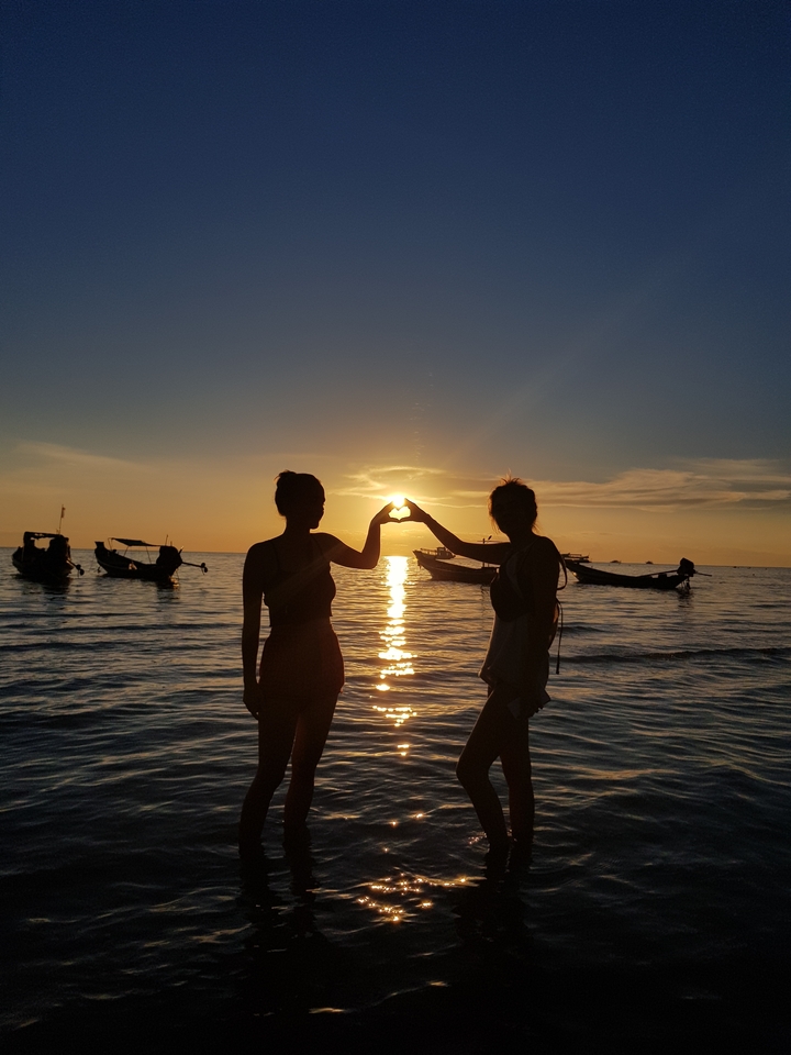 Silhouettes of people forming a heart shape at sunset with boats.