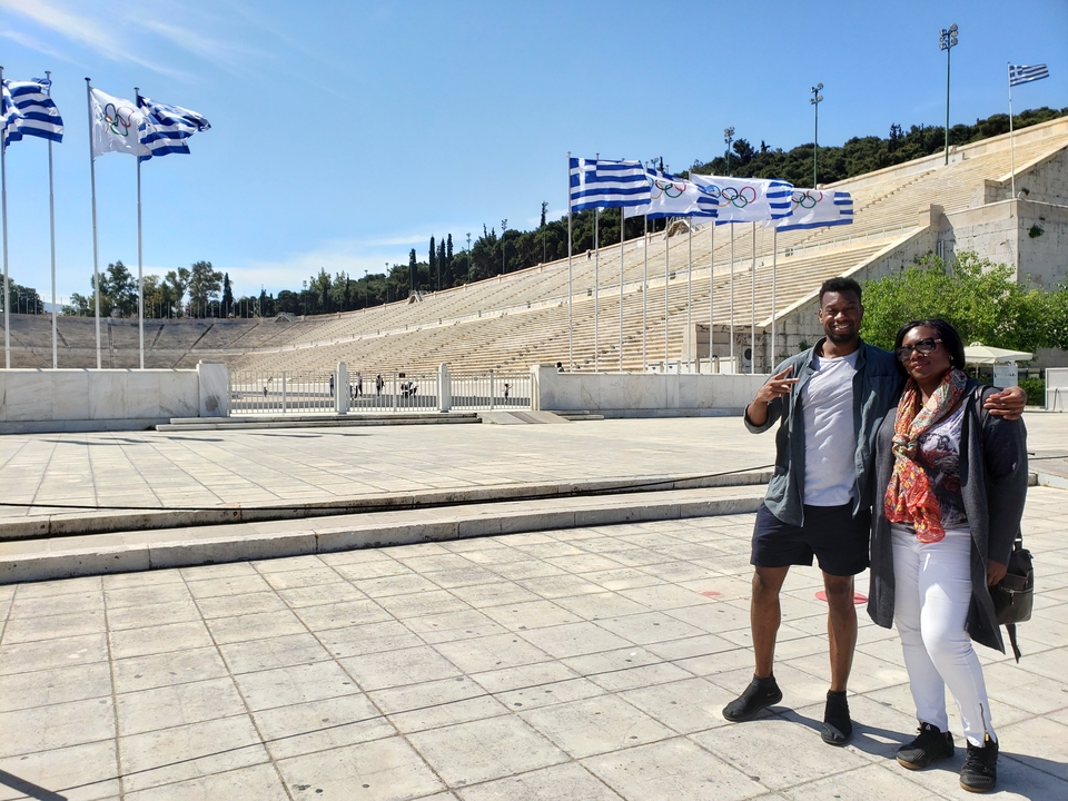Deux personnes posant devant un stade avec des drapeaux grecs.