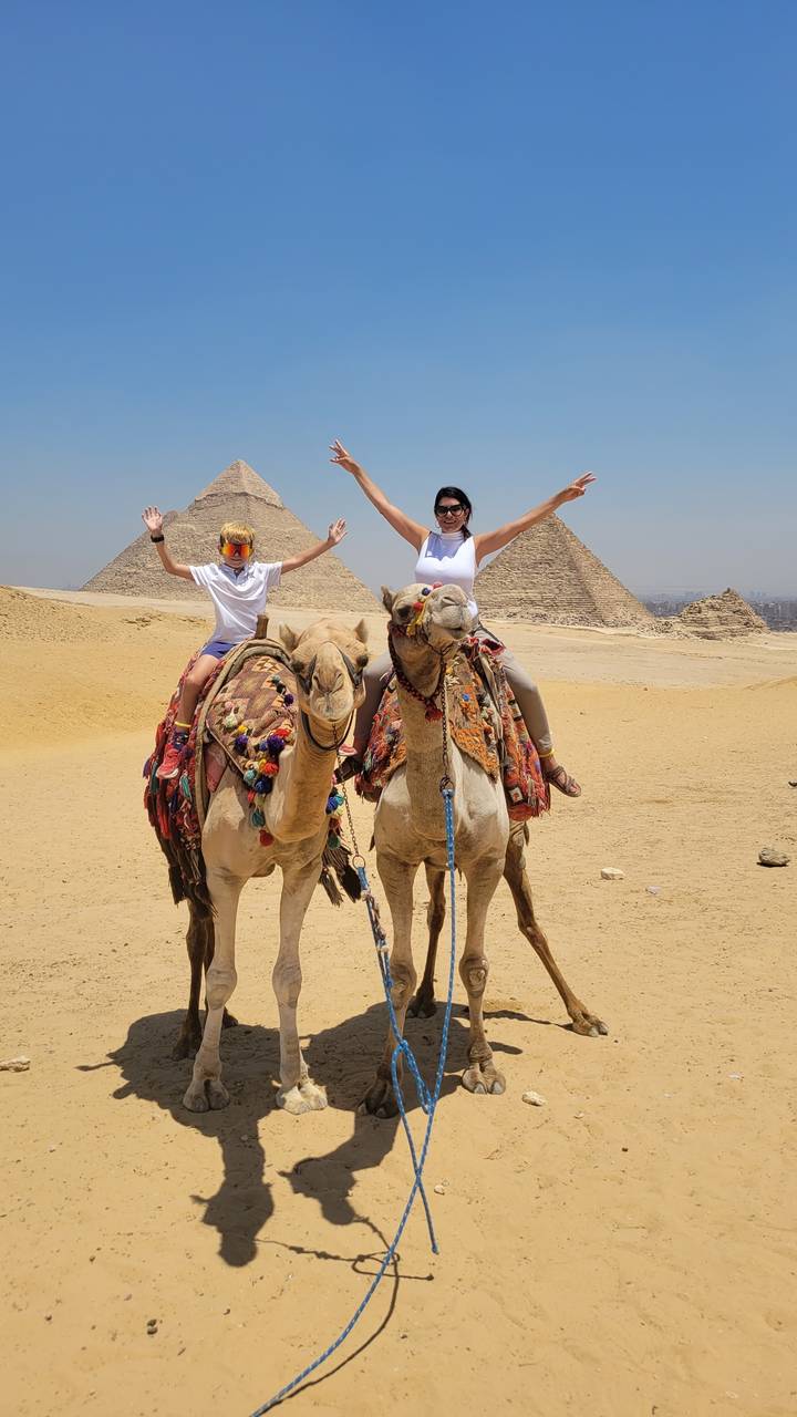 Two individuals riding camels in the desert with pyramids in the background.
