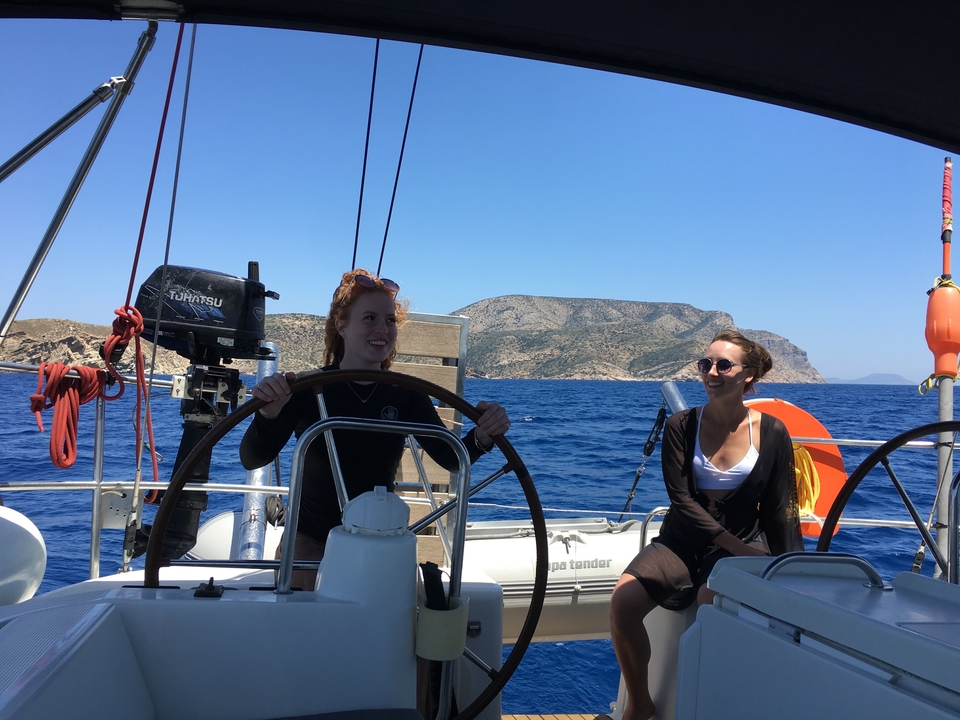 Deux personnes dirigeant un voilier avec une île rocheuse en arrière-plan.