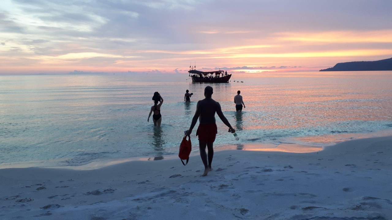People walking into the sea with a silhouette of a boat at sunset.