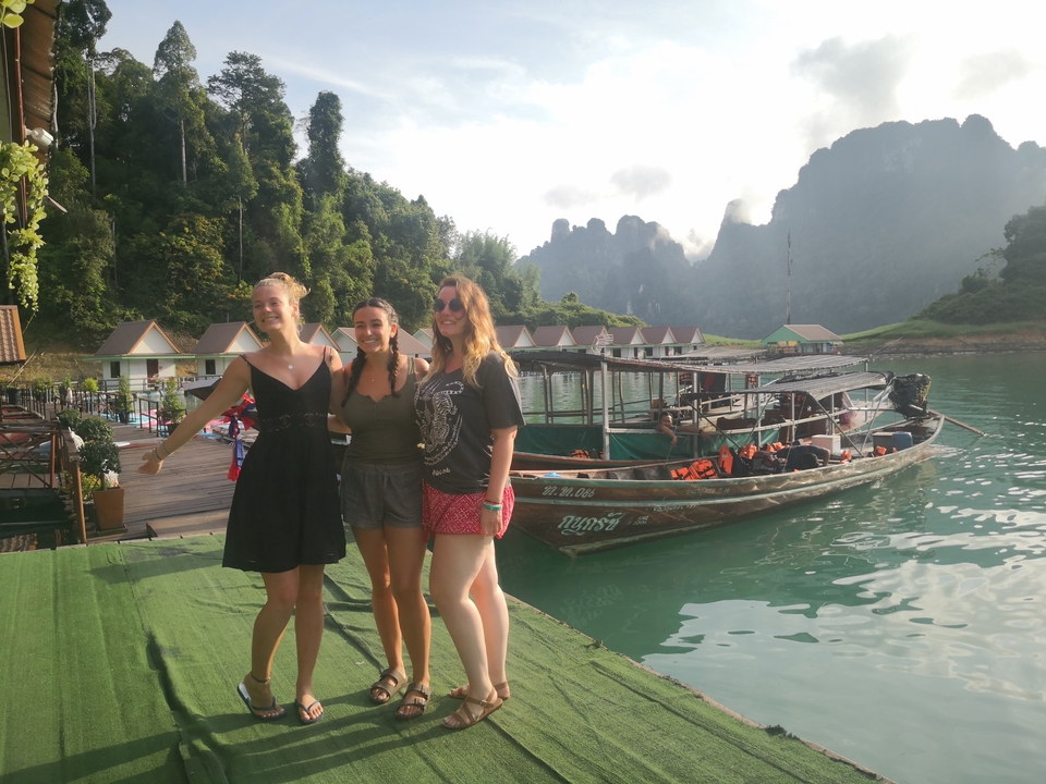 Group of people on a lakeside with boats and scenic mountainous backdrop.