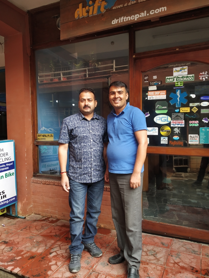 Two men pose together outside a tour office decorated with stickers and adventure posters.