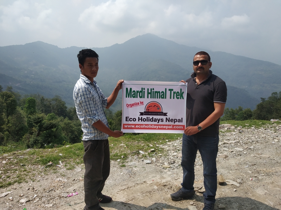 Two men hold a banner advertising a Mardi Himal trek against a hazy Himalayan backdrop.