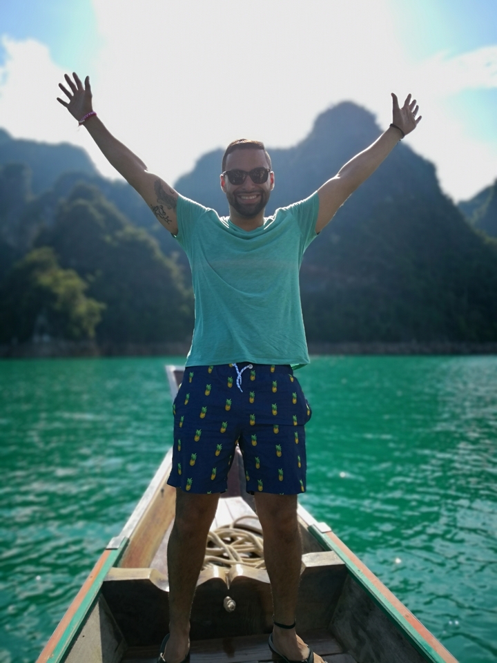 Man posing with arms raised on a boat with a scenic backdrop.