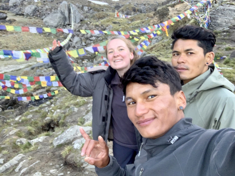 Three trekkers smile for a selfie beside colourful prayer flags in a rocky alpine landscape.