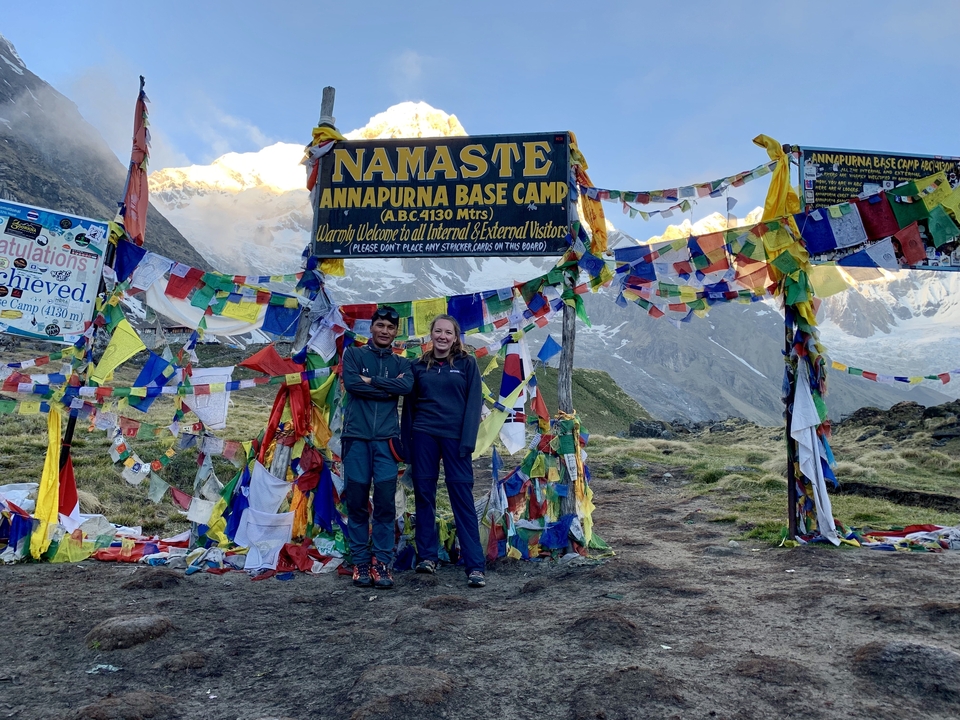 Two trekkers pose arm-in-arm beneath the Annapurna Base Camp welcome sign surrounded by bright prayer flags and snowy peaks.