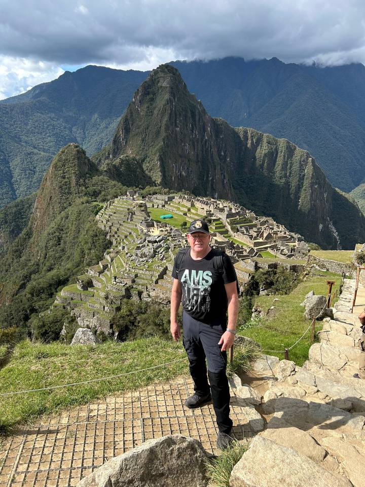 Mann posiert in Machu Picchu mit sichtbaren Ruinen und Bergen.