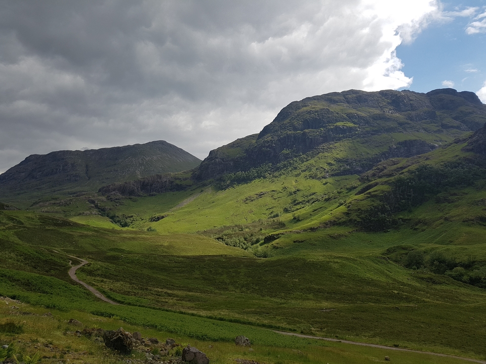 Vue panoramique de collines vertes et de montagnes rocheuses sous un ciel nuageux.