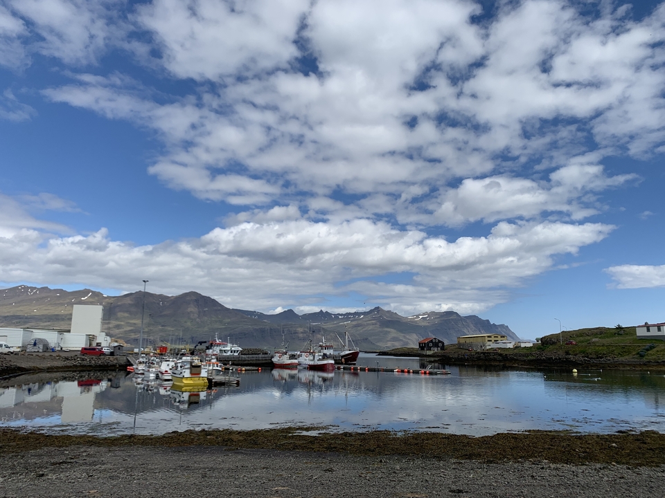 Small harbor with boats and mountains in the background under a cloudy sky.