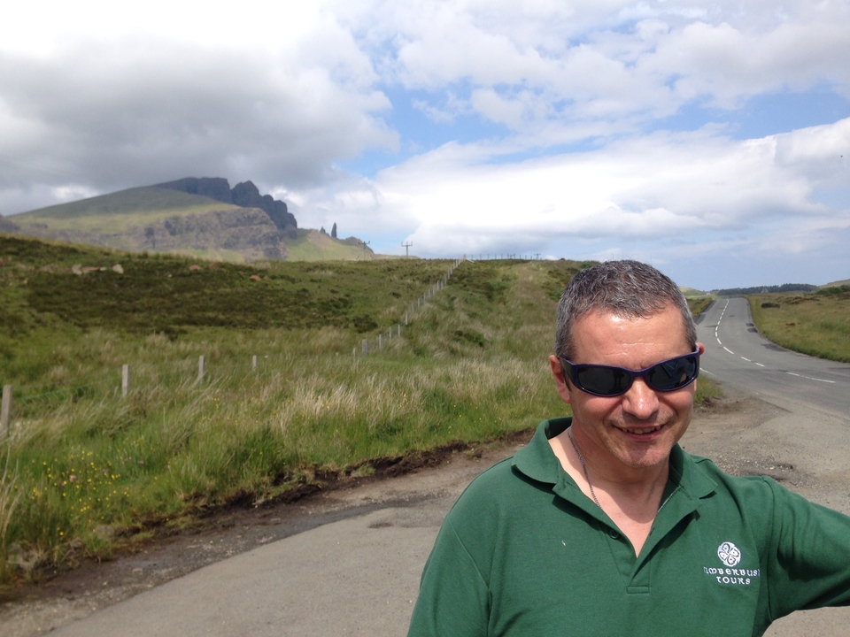 Homme souriant avec des lunettes de soleil debout sur une route avec des collines au loin.