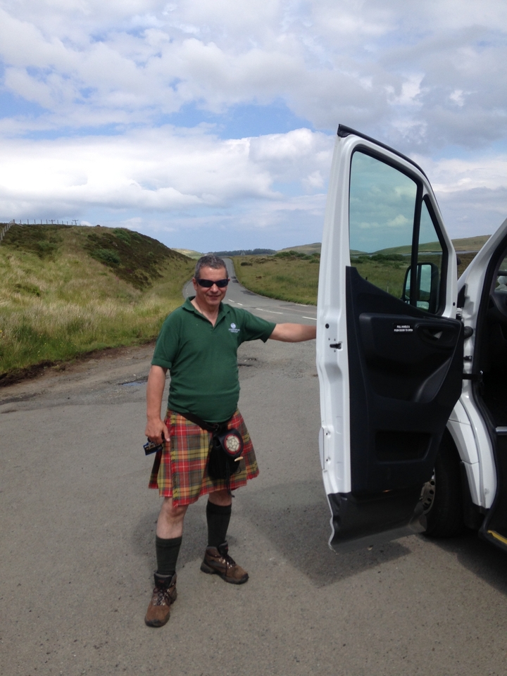 Homme en tenue traditionnelle debout près d'une portière de véhicule ouverte sur une route rurale.