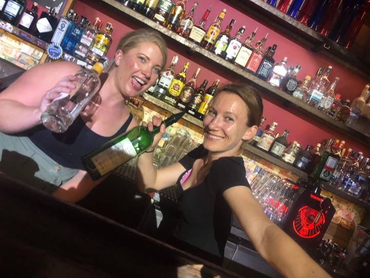 Two women behind a bar holding bottles.
