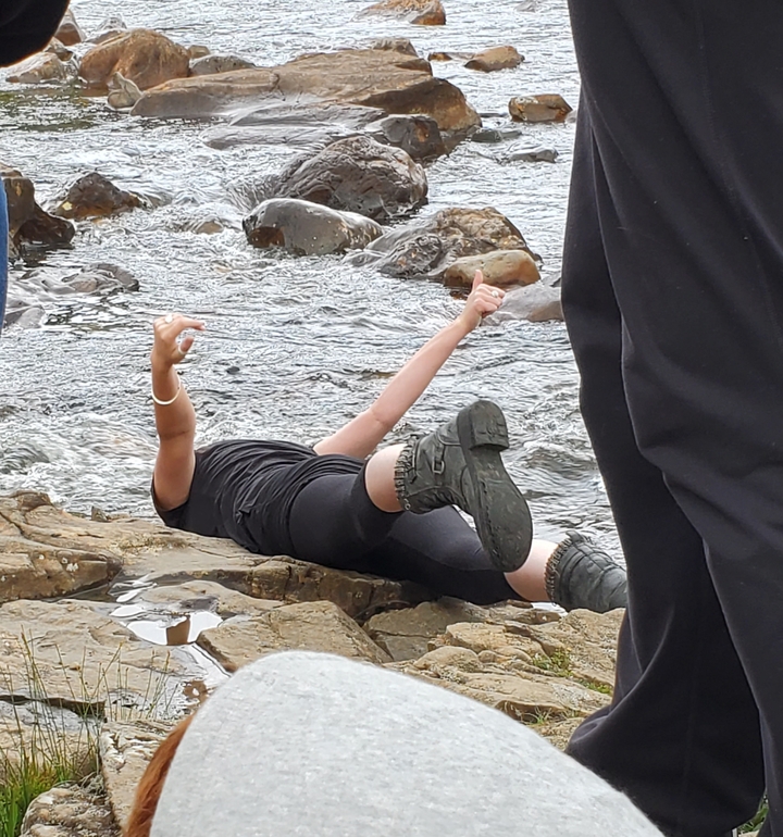 Person lying on rocks near a river, gesturing happily.