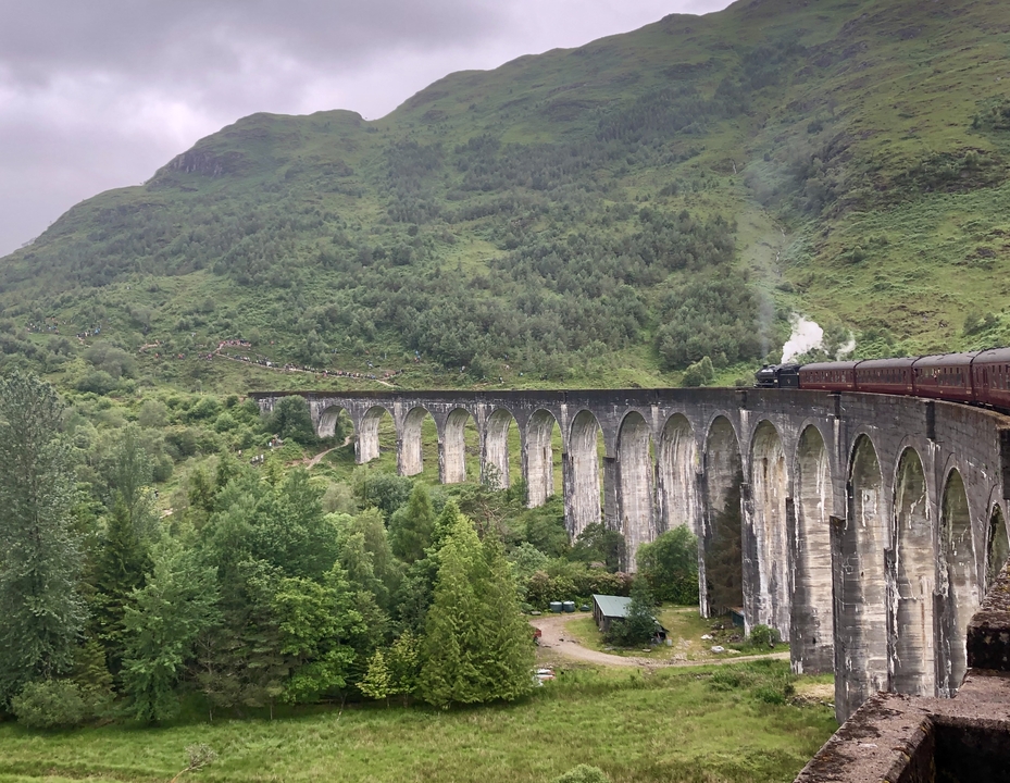 Un train traversant le viaduc emblématique de Glenfinnan avec des collines verdoyantes en arrière-plan sous un ciel nuageux.