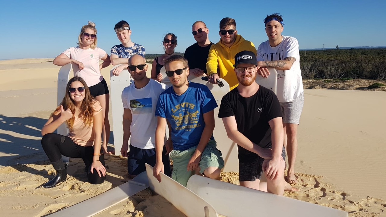 Photo de groupe de personnes posant avec des planches de surf sur une plage de sable.