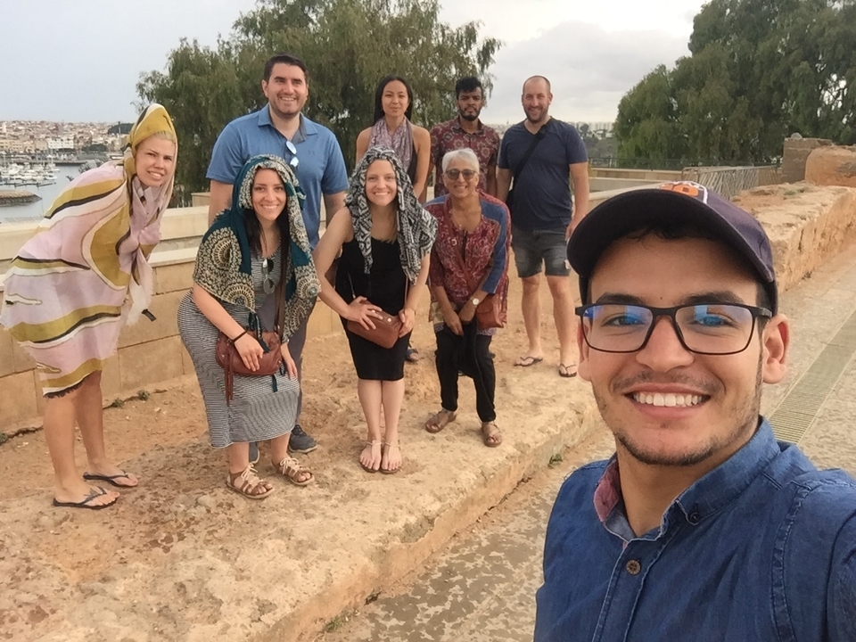 A smiling tour group poses for a selfie on a coastal rampart overlooking a marina and cityscape in Morocco.