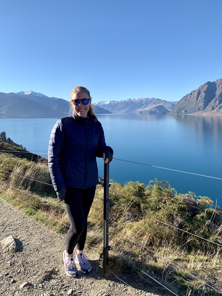Femme avec des lunettes de soleil posant près d'un lac avec des montagnes.