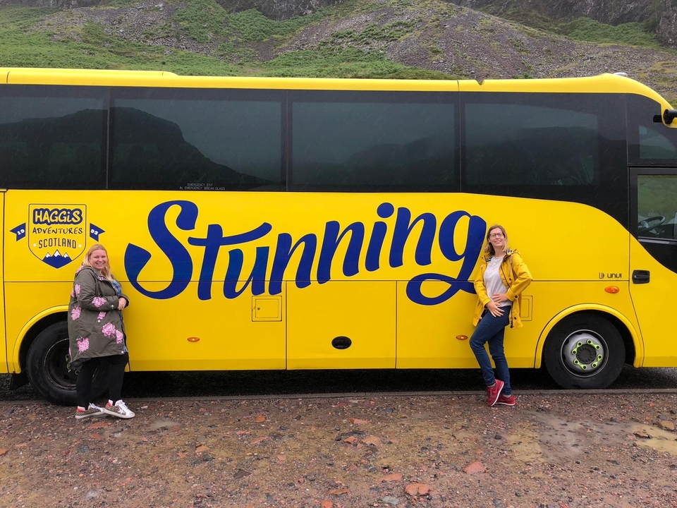 Deux personnes posent à côté d'un bus jaune avec 'Stunning' écrit dessus.