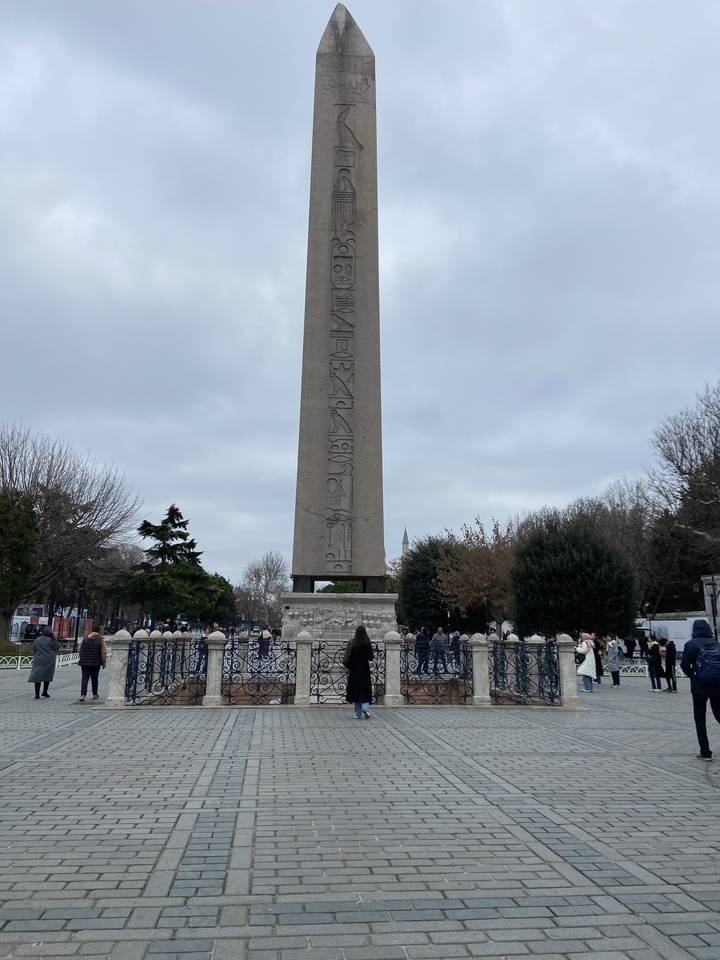 Tall stone obelisk surrounded by tourists in a park.
**French translation:**
Grand obélisque de pierre entouré de touristes dans un parc.