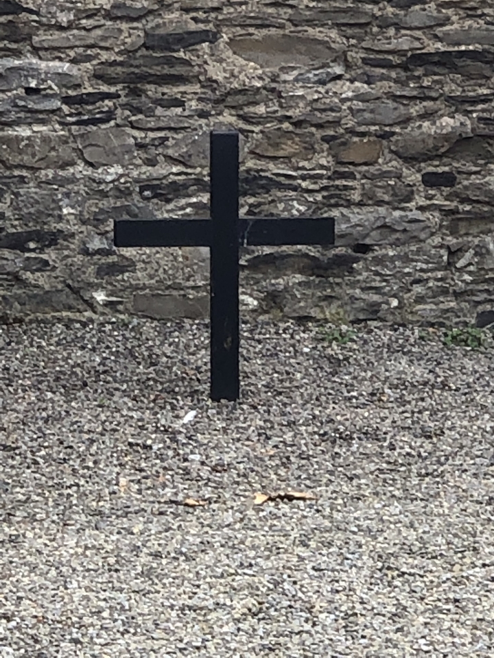 Une croix noire solitaire sur du gravier avec un mur de pierre en arrière-plan.