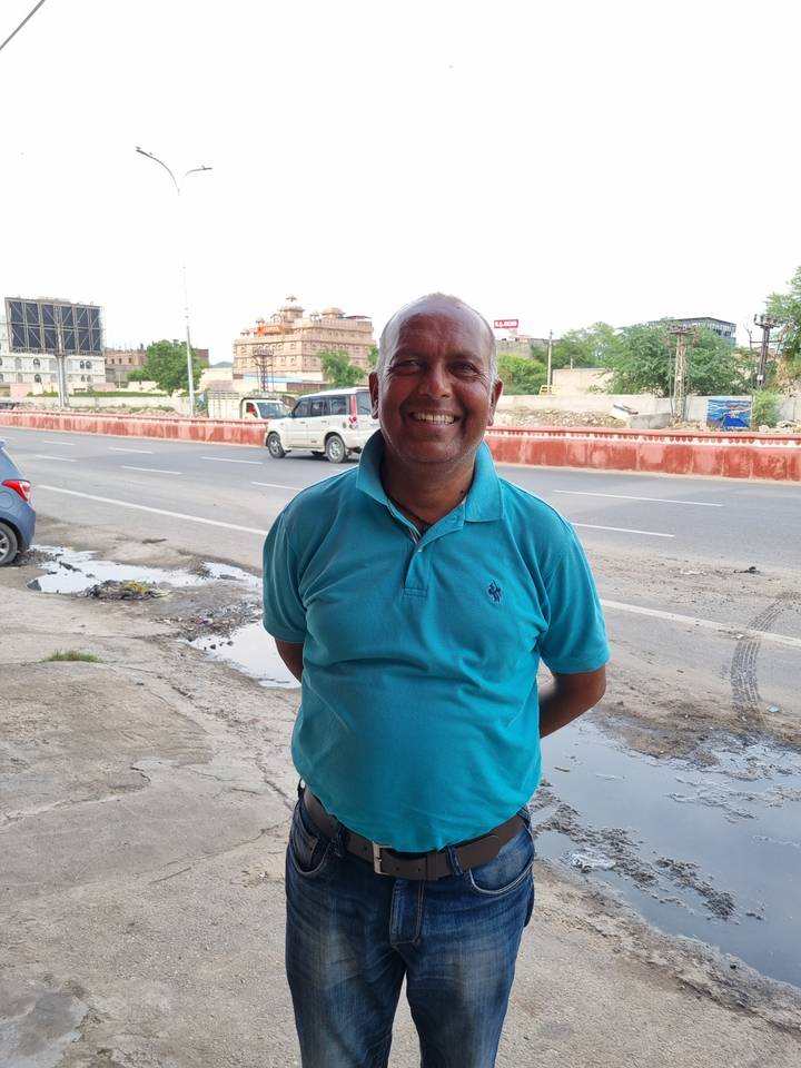 Un homme souriant en chemise bleue se tient au bord de la route avec une vue lointaine d'un bâtiment et d'une voiture garée.