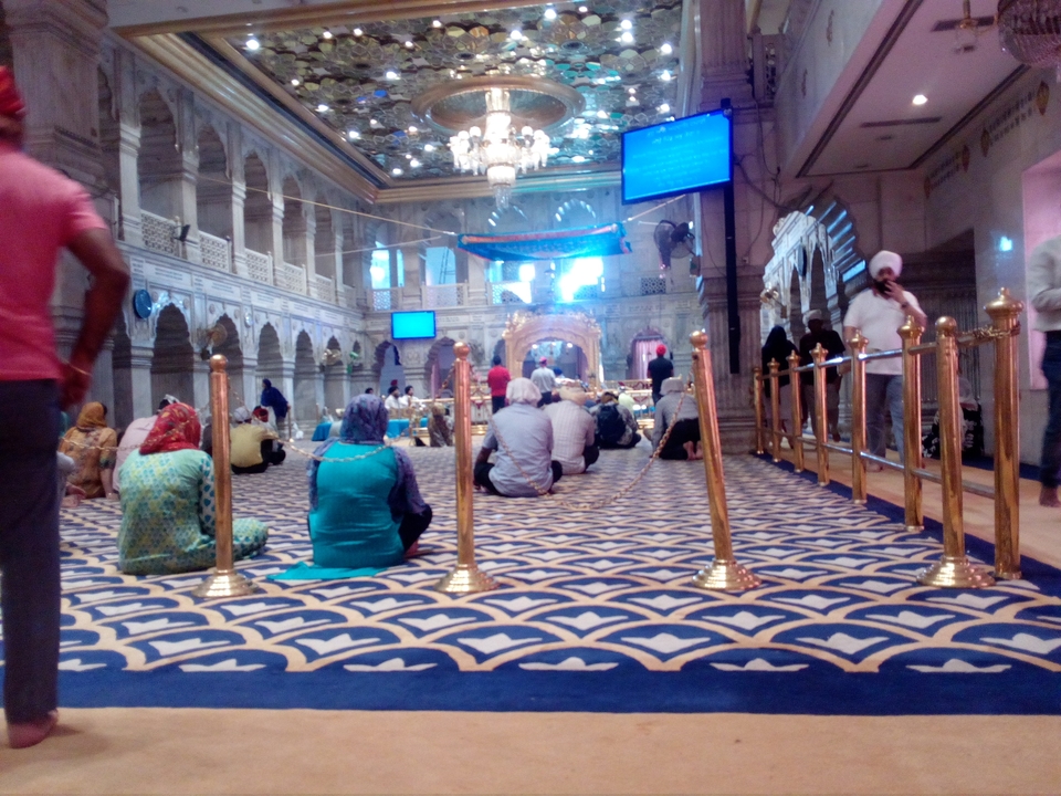 Worshippers sit cross-legged on patterned carpet inside a Sikh gurdwara, facing the ornate altar under chandeliers and screens.